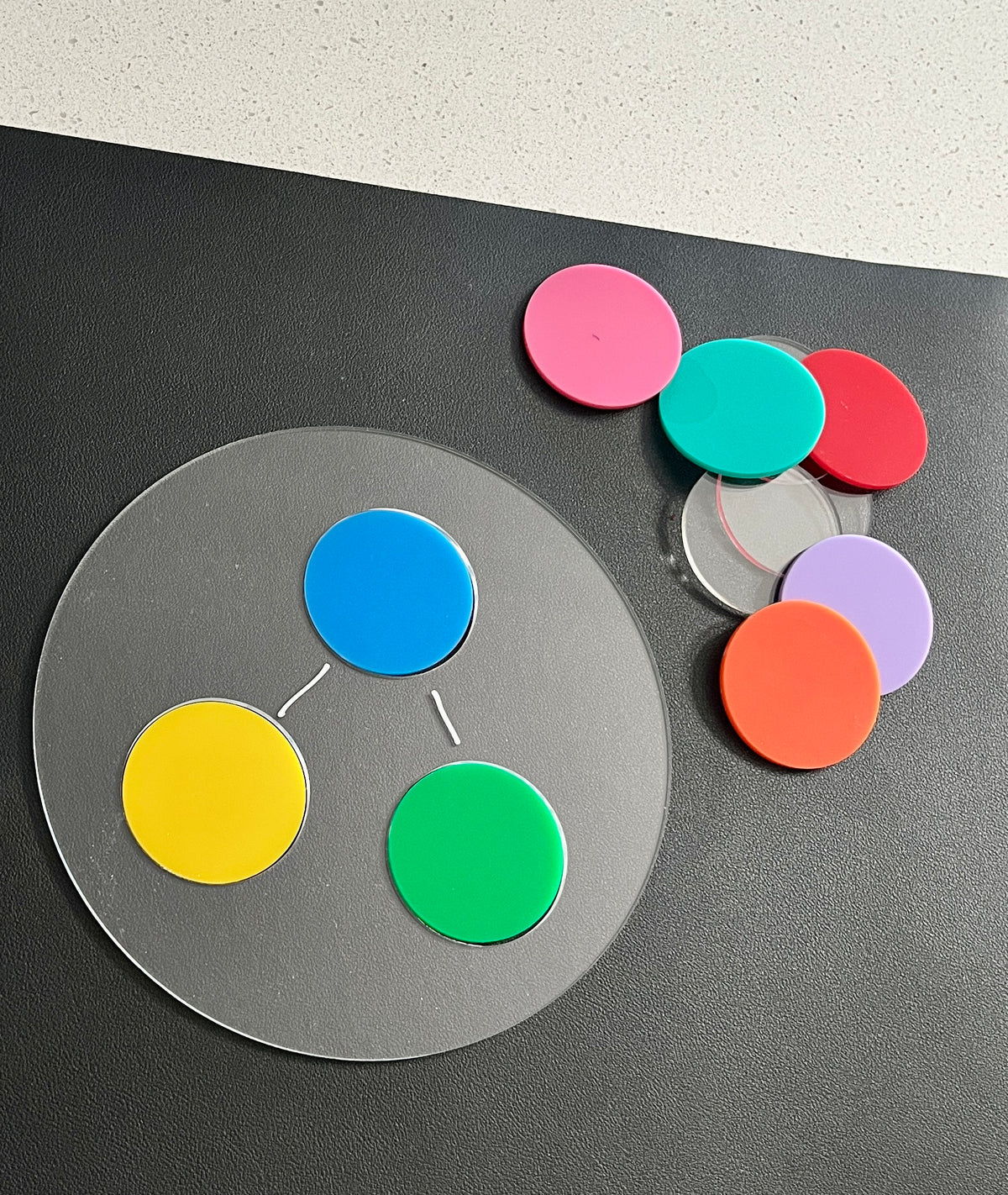 Clear circular board with connected blue, yellow, and green discs beside scattered colorful translucent discs on black surface