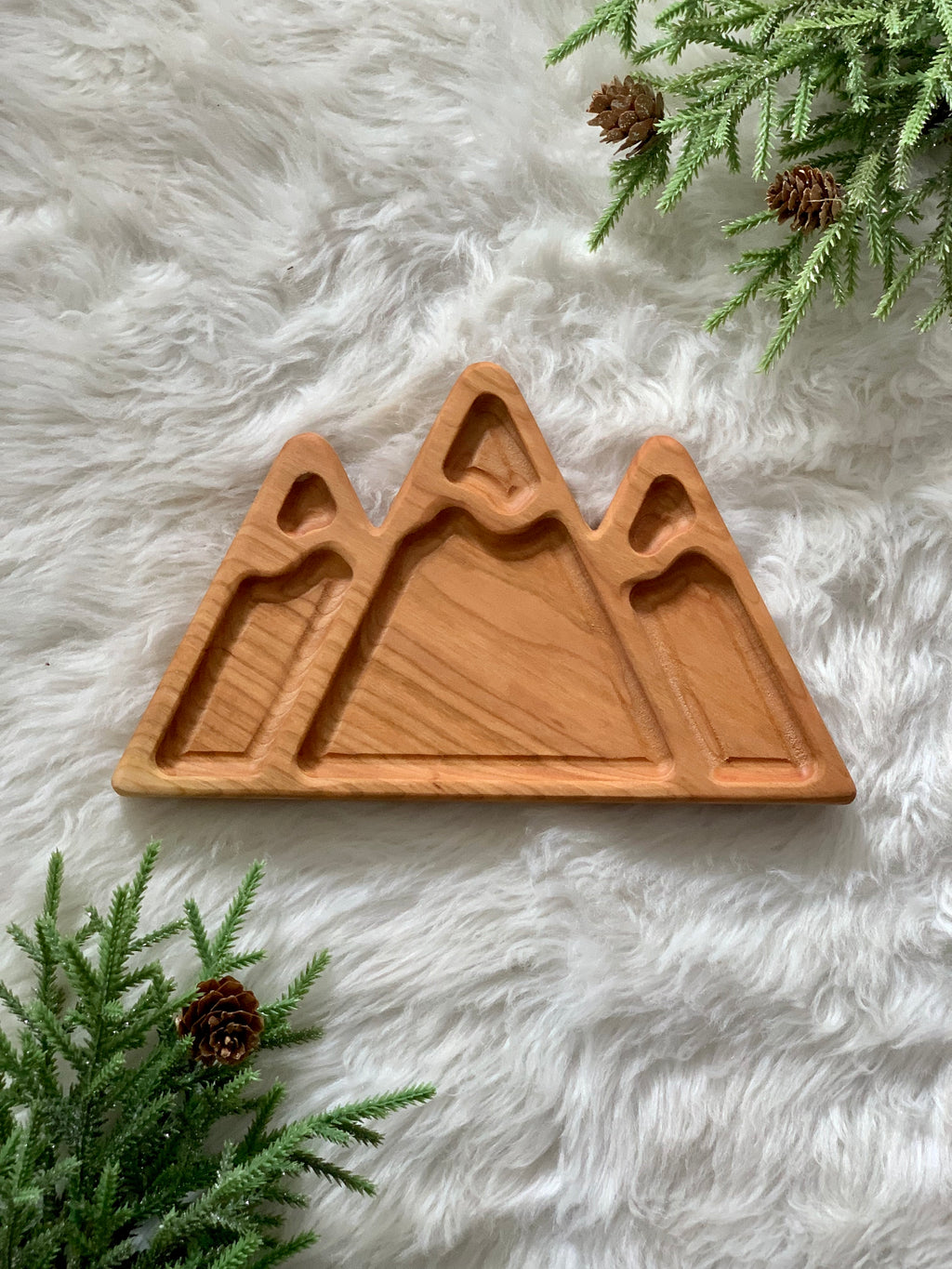 Mountain-shaped wooden sensory tray with three carved peaks on white fur background with pine branches and cones