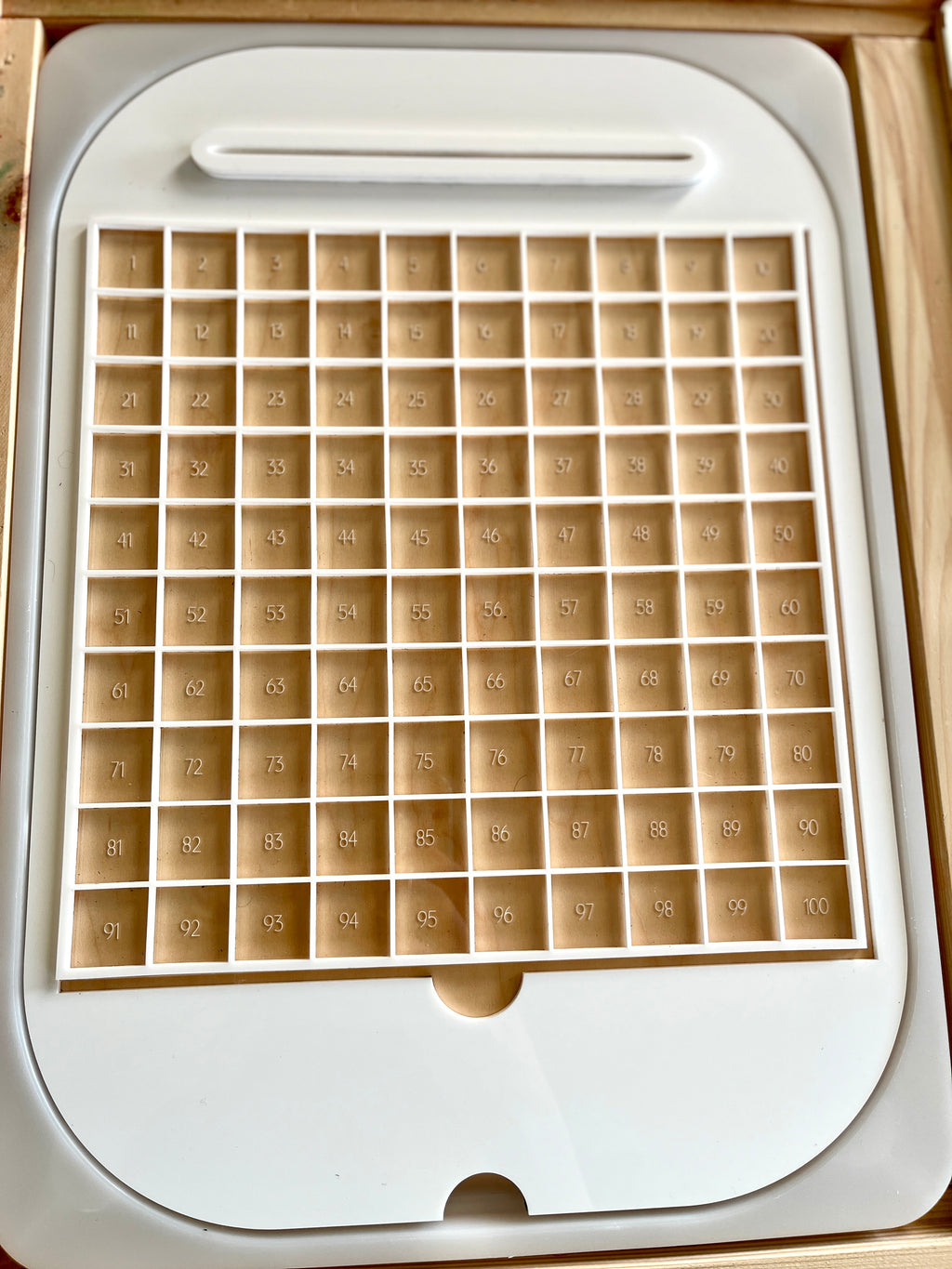 White plastic counting tray with 100 numbered square compartments arranged in a 10 by 10 grid on beige background