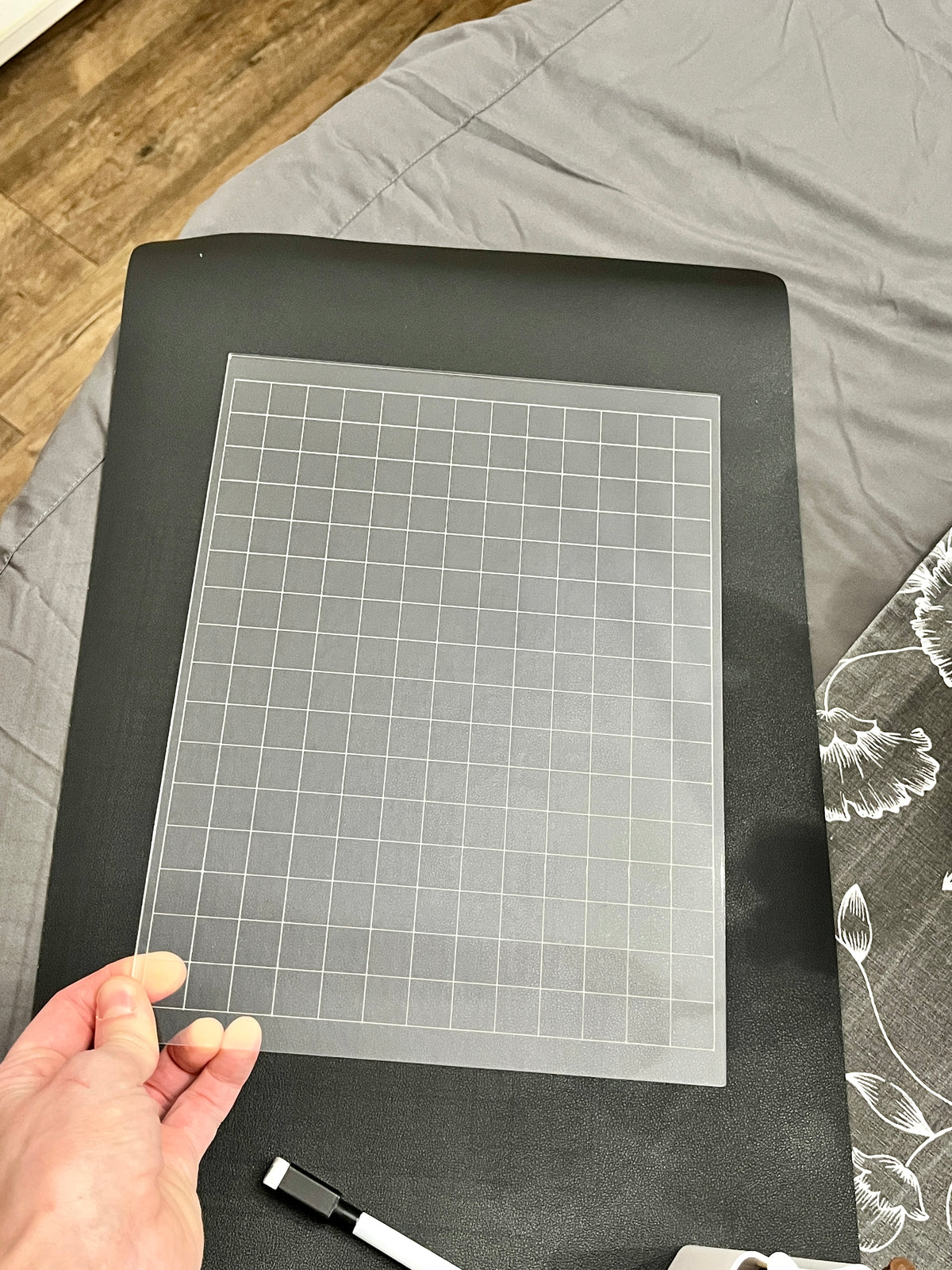 Transparent grid template with white lines held over black surface beside a black and white marker on fabric background