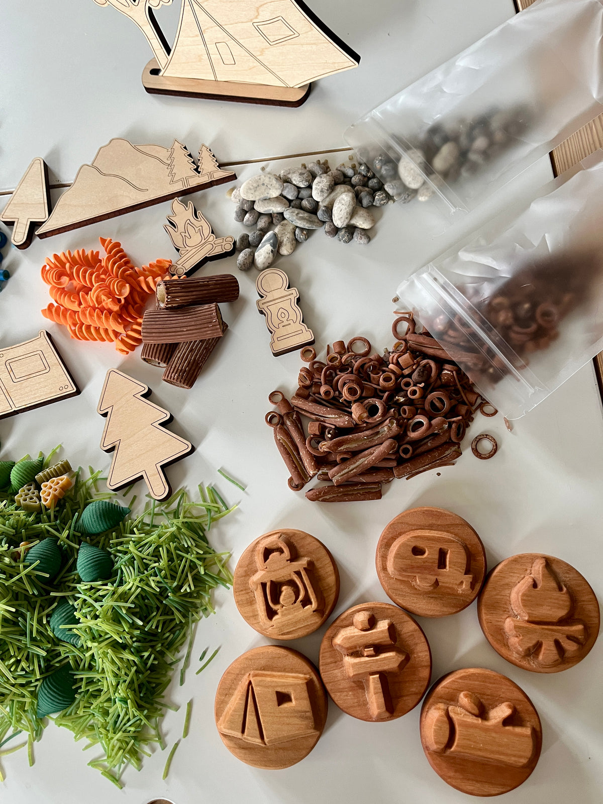 Wooden camping-themed stamps and cutouts with textured play pieces including pasta, green shredded material, pebbles, and plastic rings on white surface