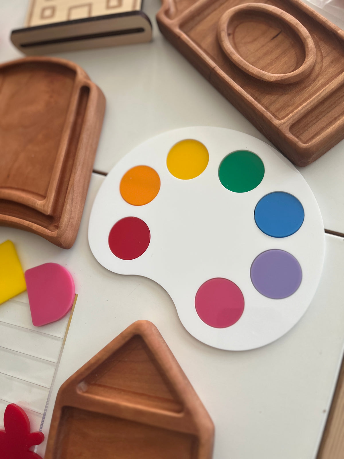 White artist palette with colorful circular paint spots surrounded by various wooden trays on white surface