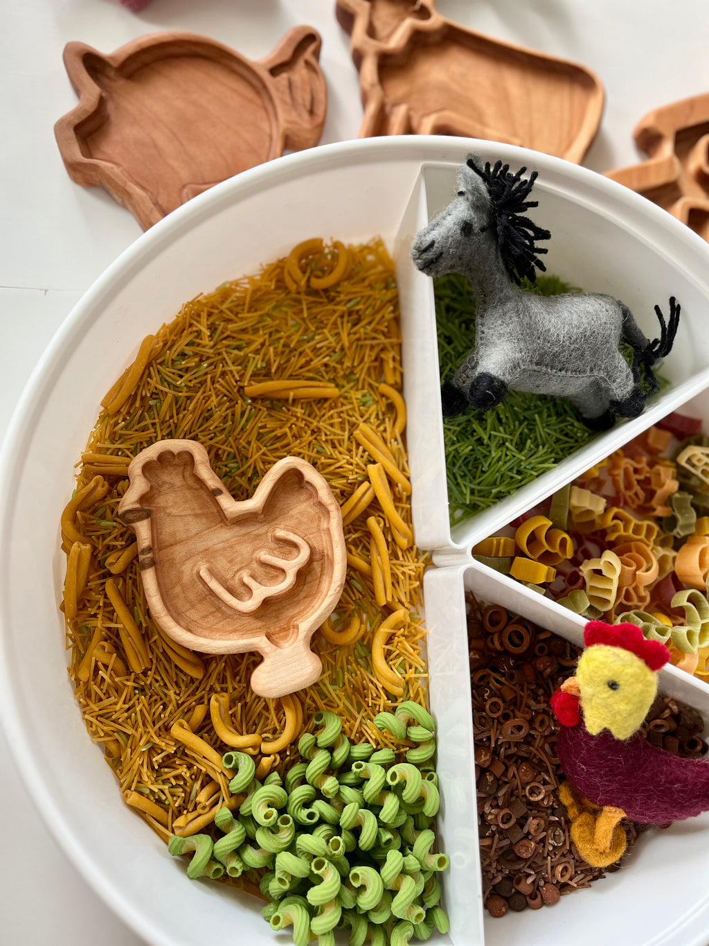 Round divided tray with colorful pasta, wooden chicken scoop, and felt donkey and chicken on white surface