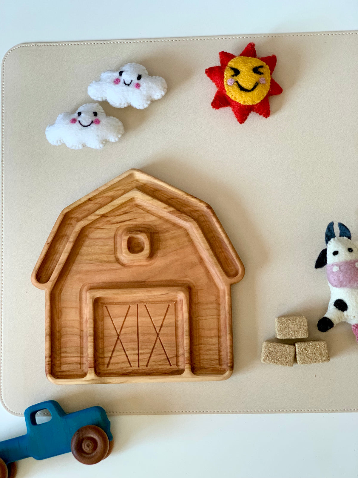 Wooden barn-shaped tray surrounded by a blue toy tractor, felt cow, hay cubes, and smiling sun and cloud decorations