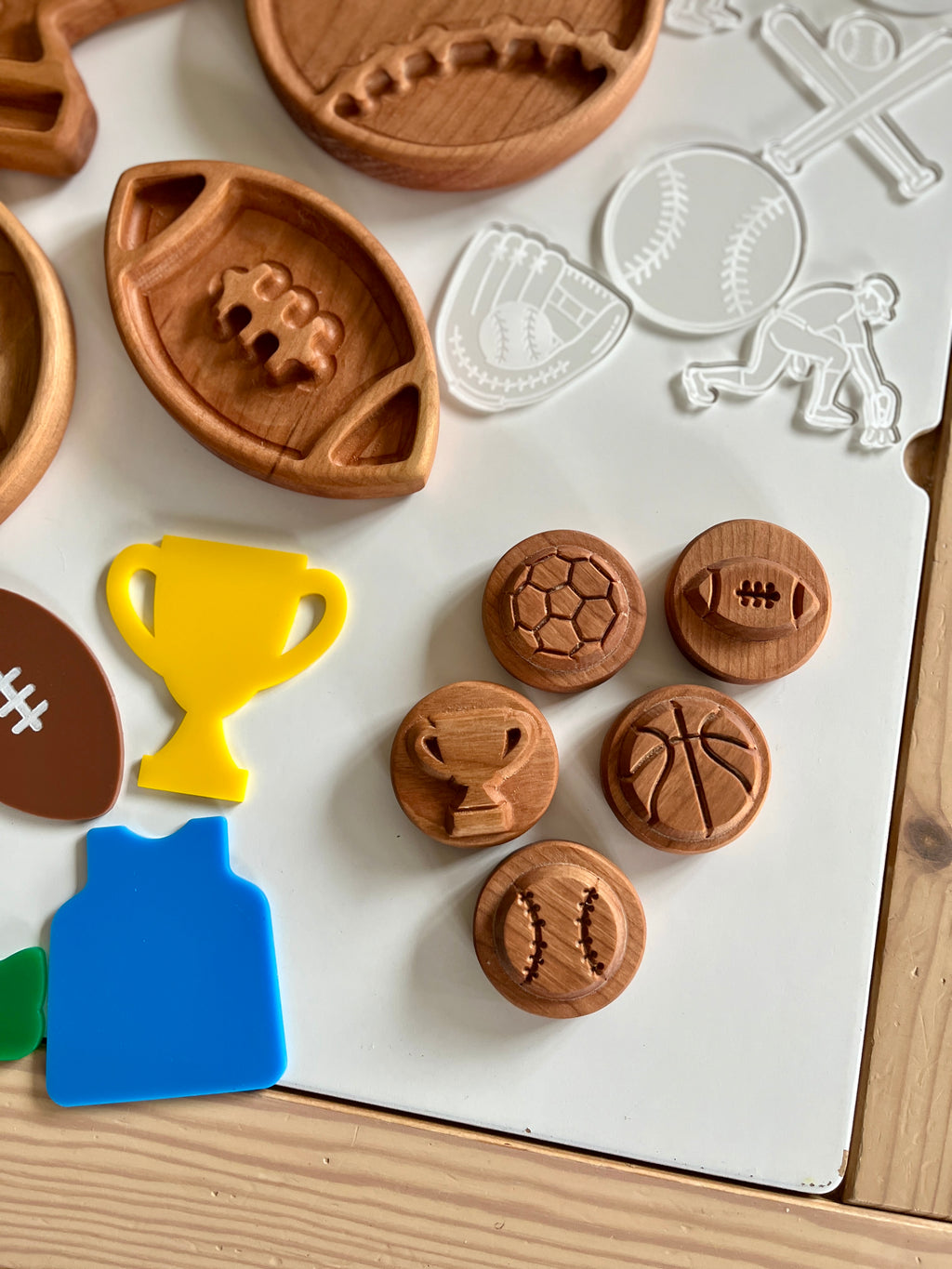 Wooden sports-themed tokens and trays featuring soccer, football, basketball, and baseball designs with colorful plastic shapes on white surface