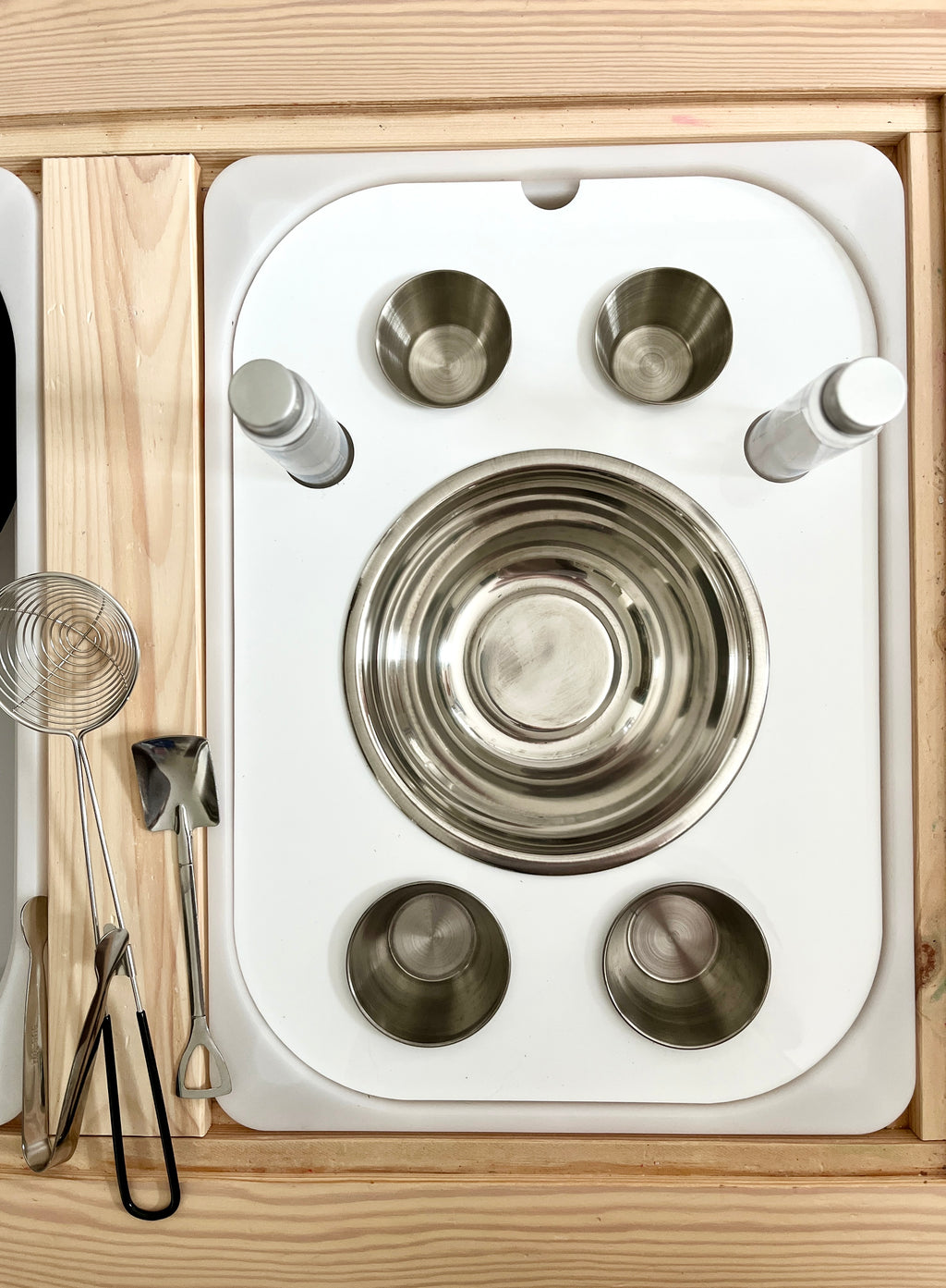 White tray with stainless steel bowls, spice shakers, and metal kitchen utensils on a wooden surface