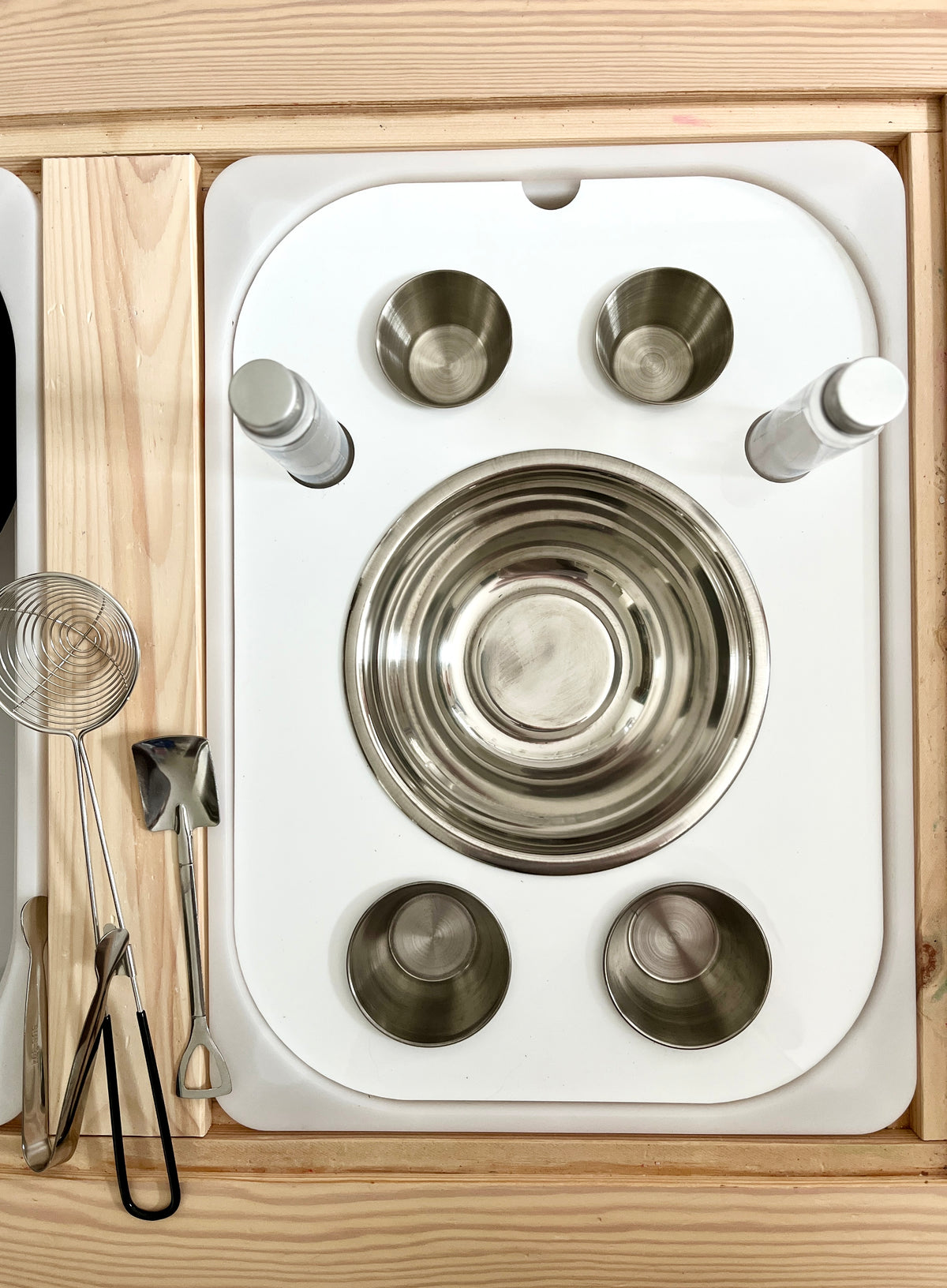 White tray with stainless steel bowls, spice shakers, and metal kitchen utensils on a wooden surface