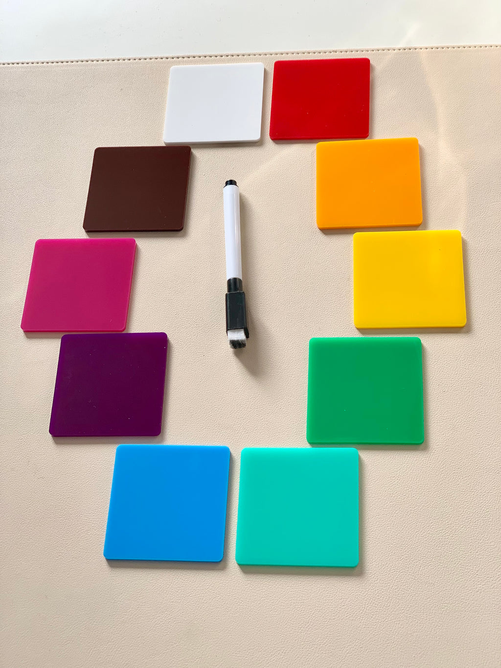 Twelve colorful square acrylic tiles arranged in a circle around a black and white dry erase marker on beige surface