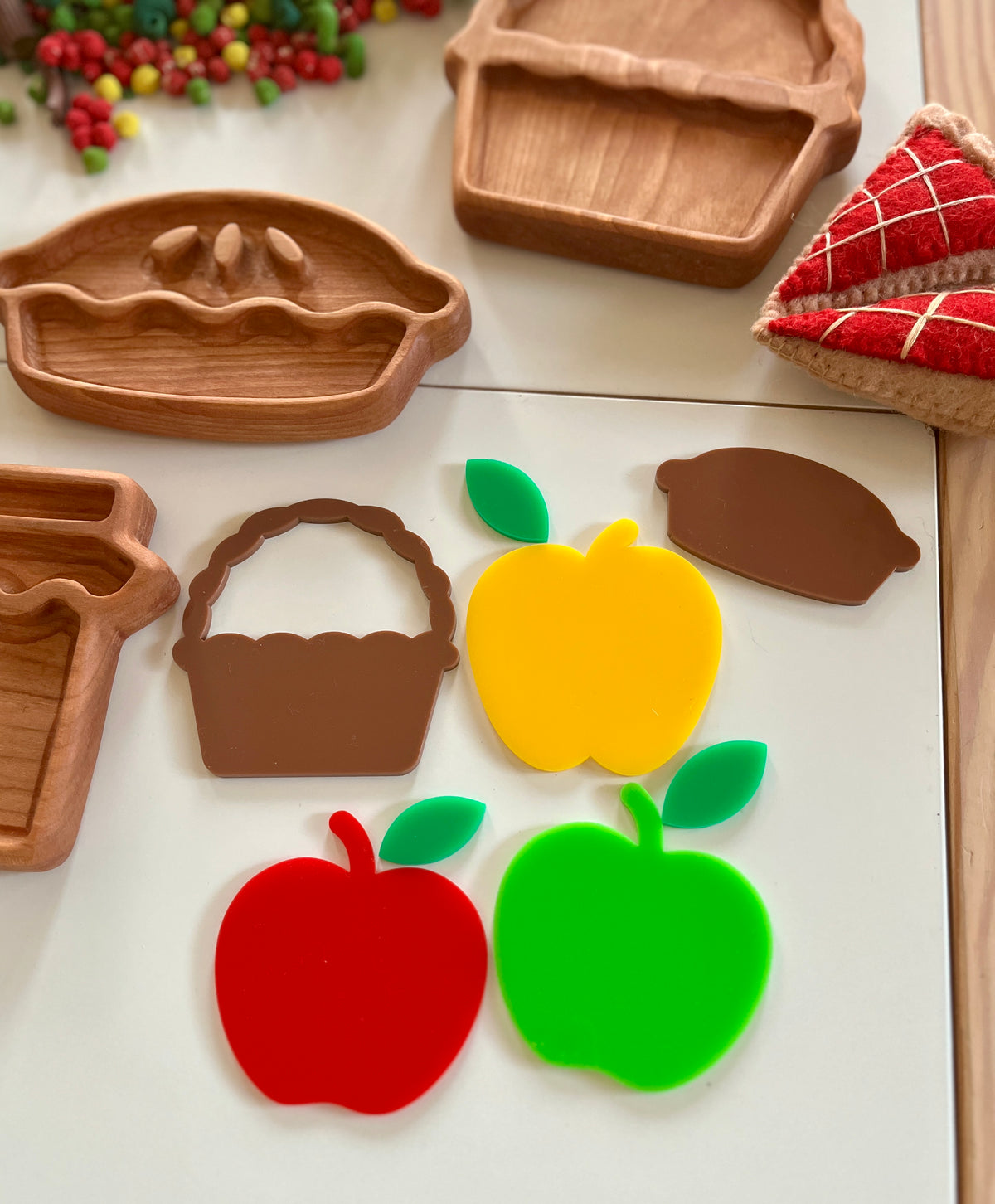 Wooden pie and basket trays with colorful plastic fruit and vegetable shapes and felt pie slices on a white surface