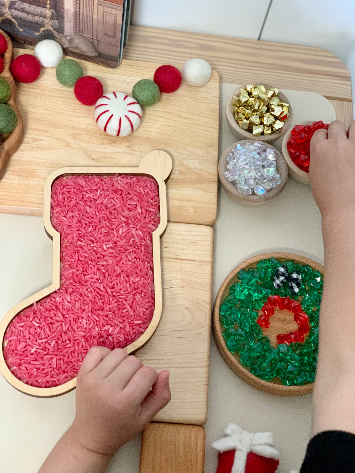 Laser cut wooden stocking sensory tray filled with red rice surrounded by festive decorations and child's hands