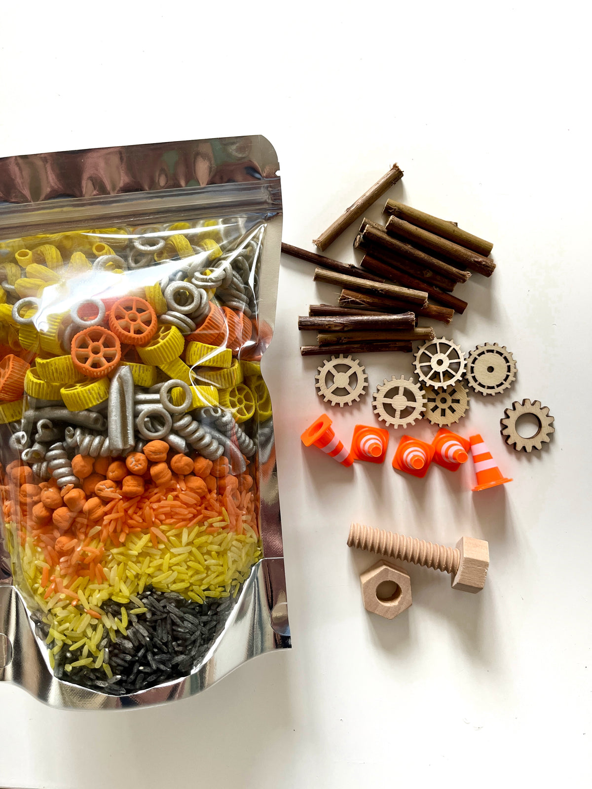 Clear pouch with multi-colored pasta, wooden twigs, gears, nuts, wooden screws, and orange traffic cones on white surface