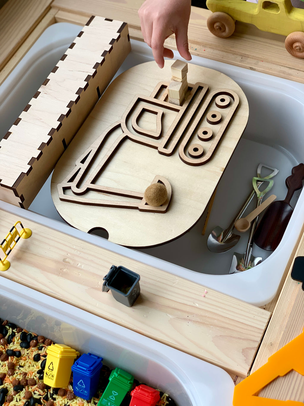 Child’s hand manipulating wooden block maze toy with small gardening tools and a yellow wooden toy truck nearby