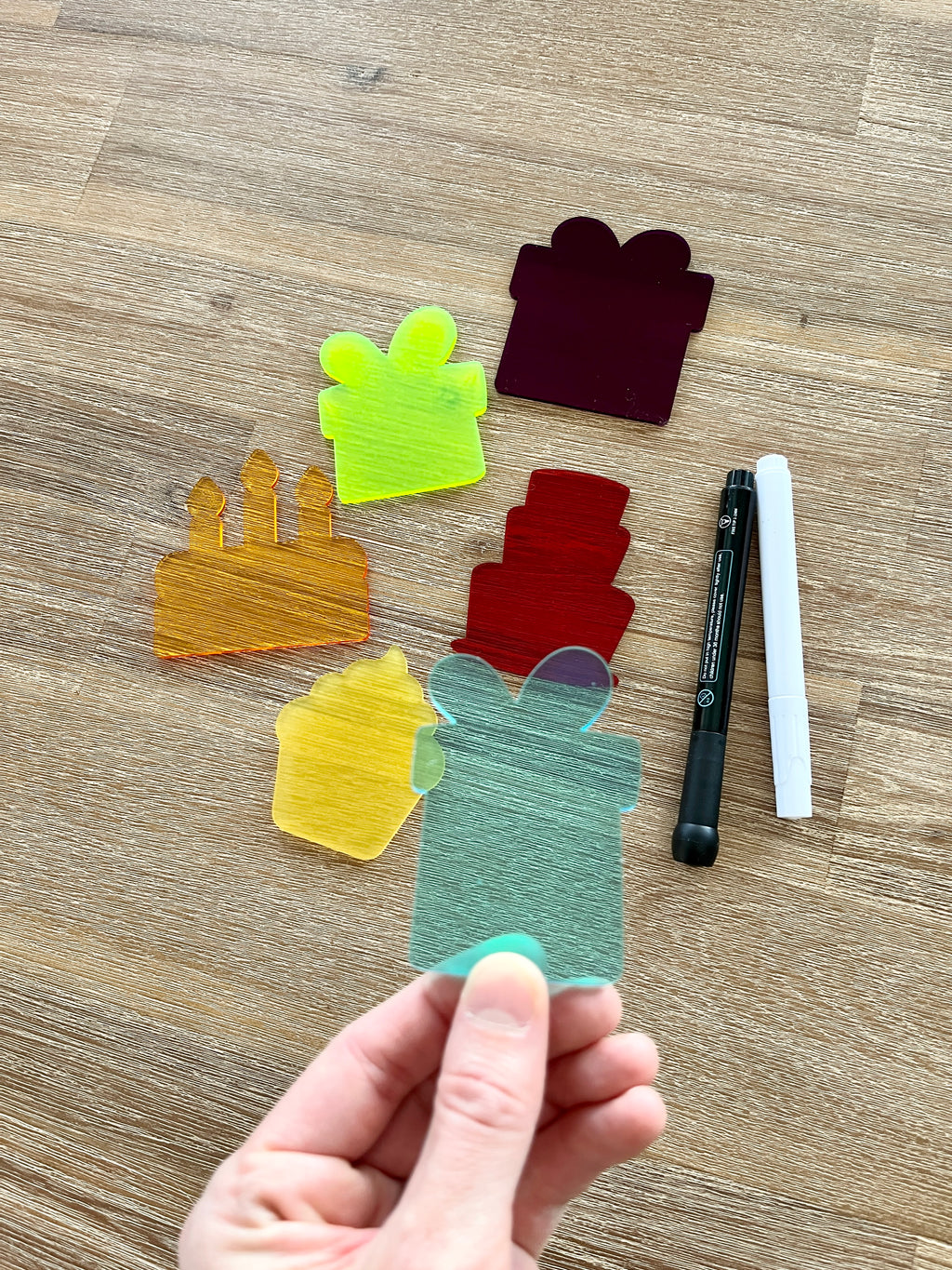 Hand holding a translucent teal gift-shaped piece with five additional colorful birthday-themed pieces and black and white dry erase markers on a wooden surface, featuring Little Dry Erasables - Translucent Birthday Set of 6, handcrafted homeschool education resources.