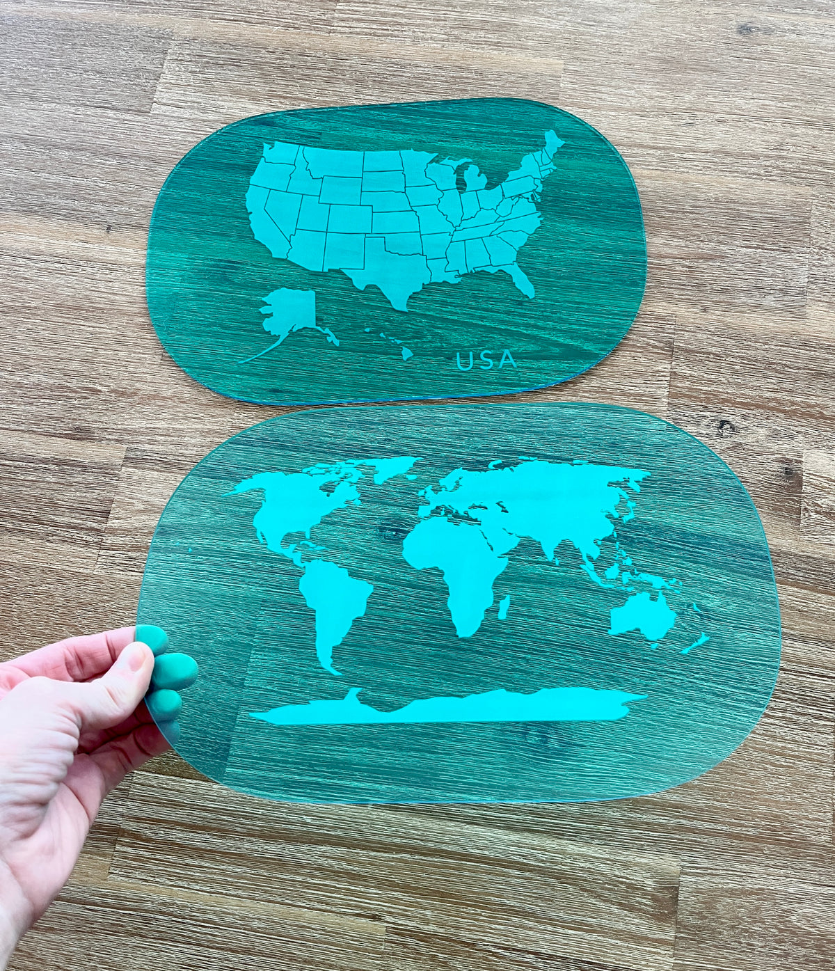Hand holding two translucent blue dry erase acrylic tracing boards featuring maps of the USA and world, showcasing Translucent Blue Maps (USA, World, Canada) Dry Erase Acrylic Tracing Board; also fits as a Flisat Insert with handcrafted, homeschool, education resources.