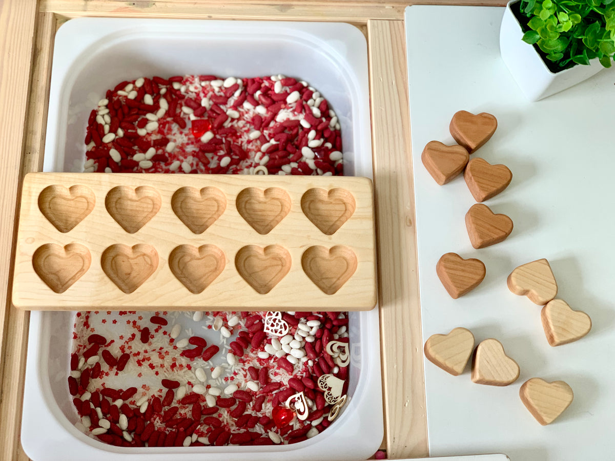 Wooden heart-shaped molds and solid wooden hearts next to a white bin filled with red and white sensory filler beads