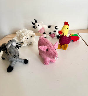 Handmade felt animal dolls including a gray donkey, white sheep, pink pig, black and white cow, and colorful rooster on a table