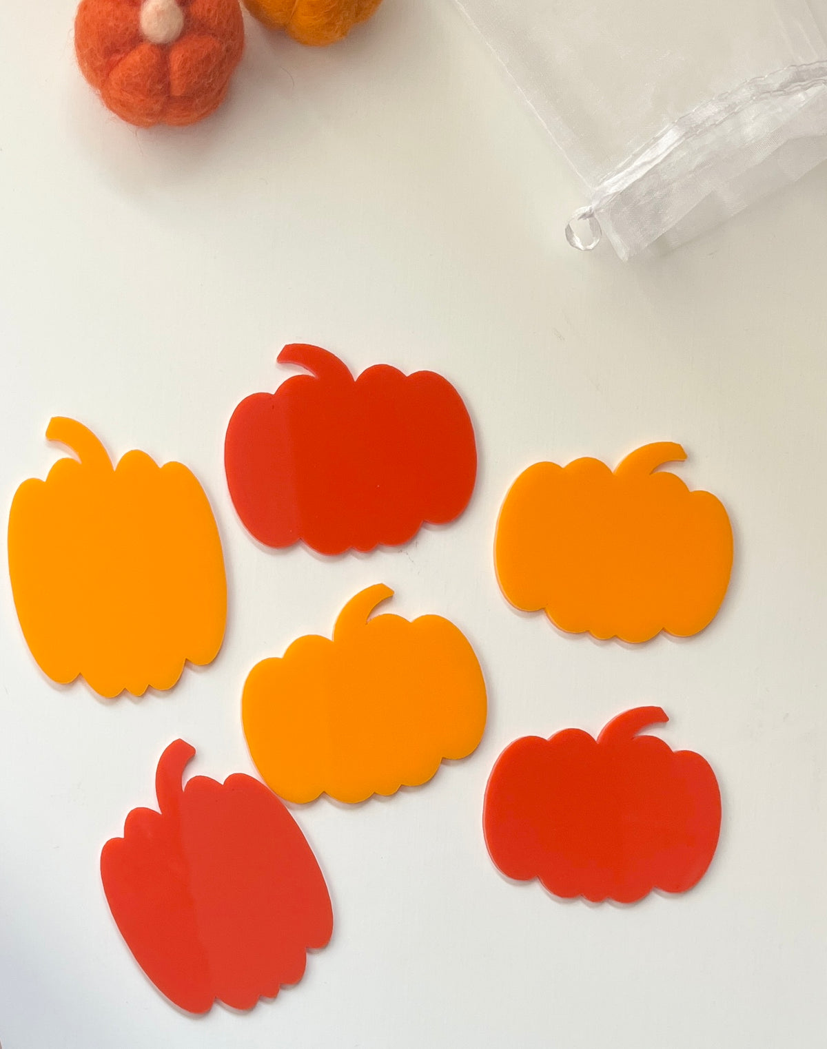 Bright orange and red pumpkin-shaped cutouts on a white surface beside a sheer white drawstring bag