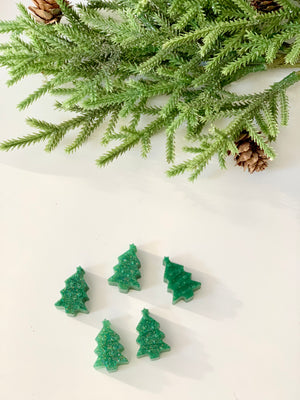 Five small green Christmas tree-shaped game pieces arranged below pine branches with cones on white surface