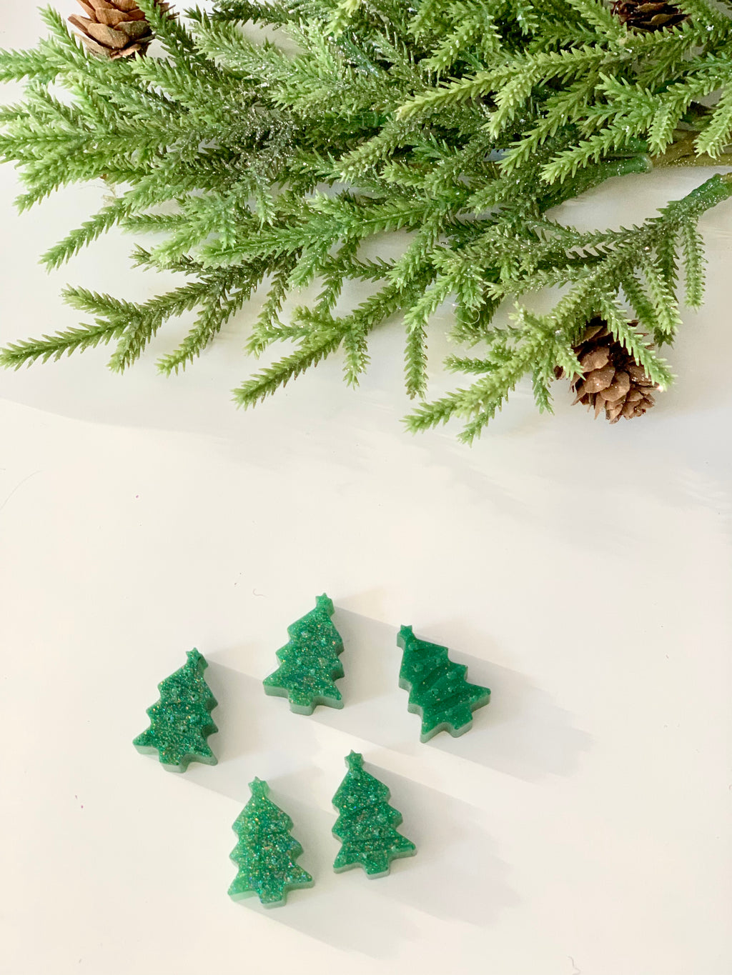 Five small green Christmas tree-shaped game pieces arranged below pine branches with cones on white surface