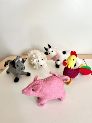 Five handmade felt animal dolls including a pink pig, multicolored rooster, black and white cow, white sheep, and gray donkey on a light surface