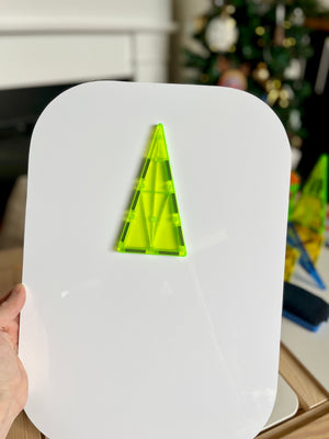 Hand holding white magnetic board with neon green translucent triangular tiles arranged in tree shape against blurred festive background