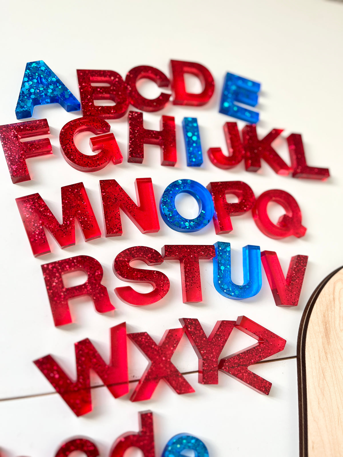 Uppercase resin alphabet letters in red and blue with glitter scattered on a white surface near a wooden edge