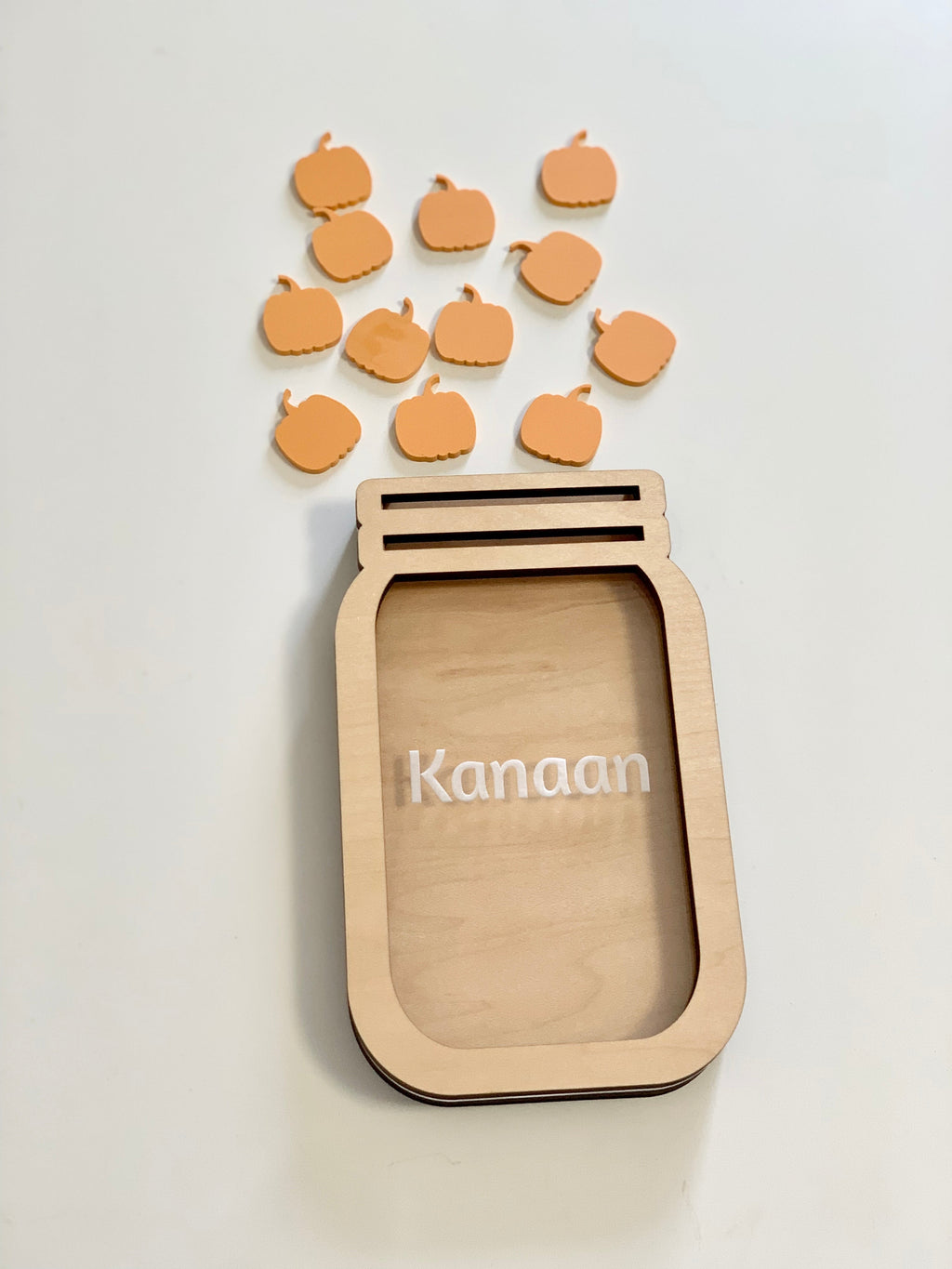 Light wooden jar-shaped frame with the name Kanaan and small orange pumpkin tokens scattered above on a white surface