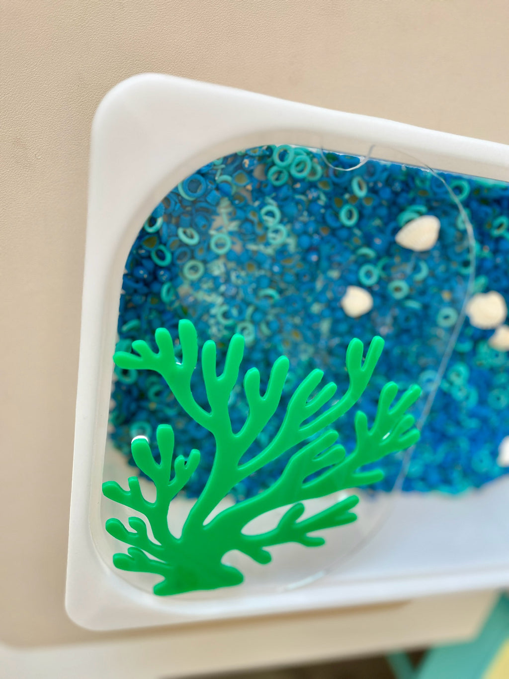 White tray with translucent cover holding blue sensory beads, small white shells, and green coral-shaped plastic decoration