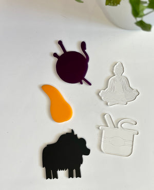 Purple snail, orange paint splatter, clear meditating figure, clear tea cup, and black buffalo acrylic shapes on white surface with plant nearby