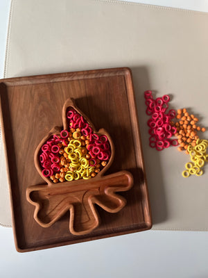 Wooden tray shaped like a flame filled with red, yellow, and orange plastic rings and beads on a beige surface