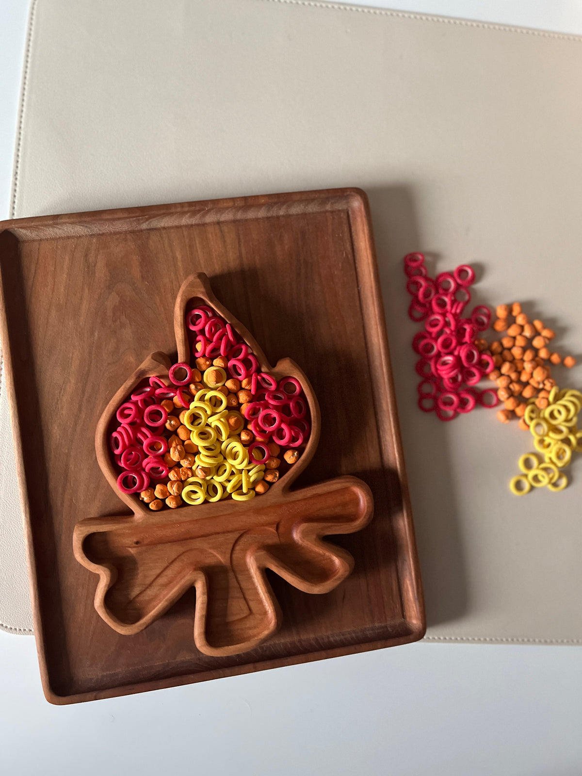 Wooden tray shaped like a flame filled with red, yellow, and orange plastic rings and beads on a beige surface
