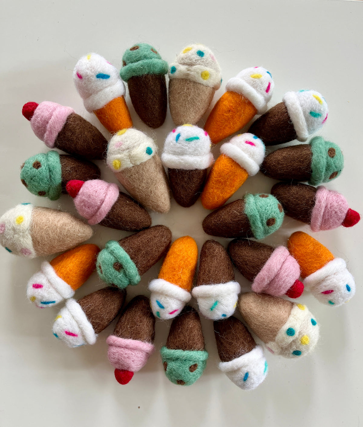 Assorted colorful felt ice cream cones with sprinkles and cherries arranged in a circular pattern on white background