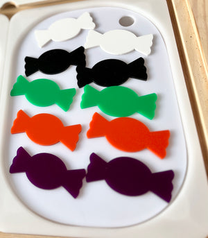 Eight candy-shaped cutouts in white, black, green, orange, and purple arranged in four rows on a white tray