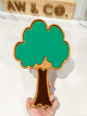 Hand holding a wooden tray shaped like a tree with green leafy top and brown trunk on a white surface