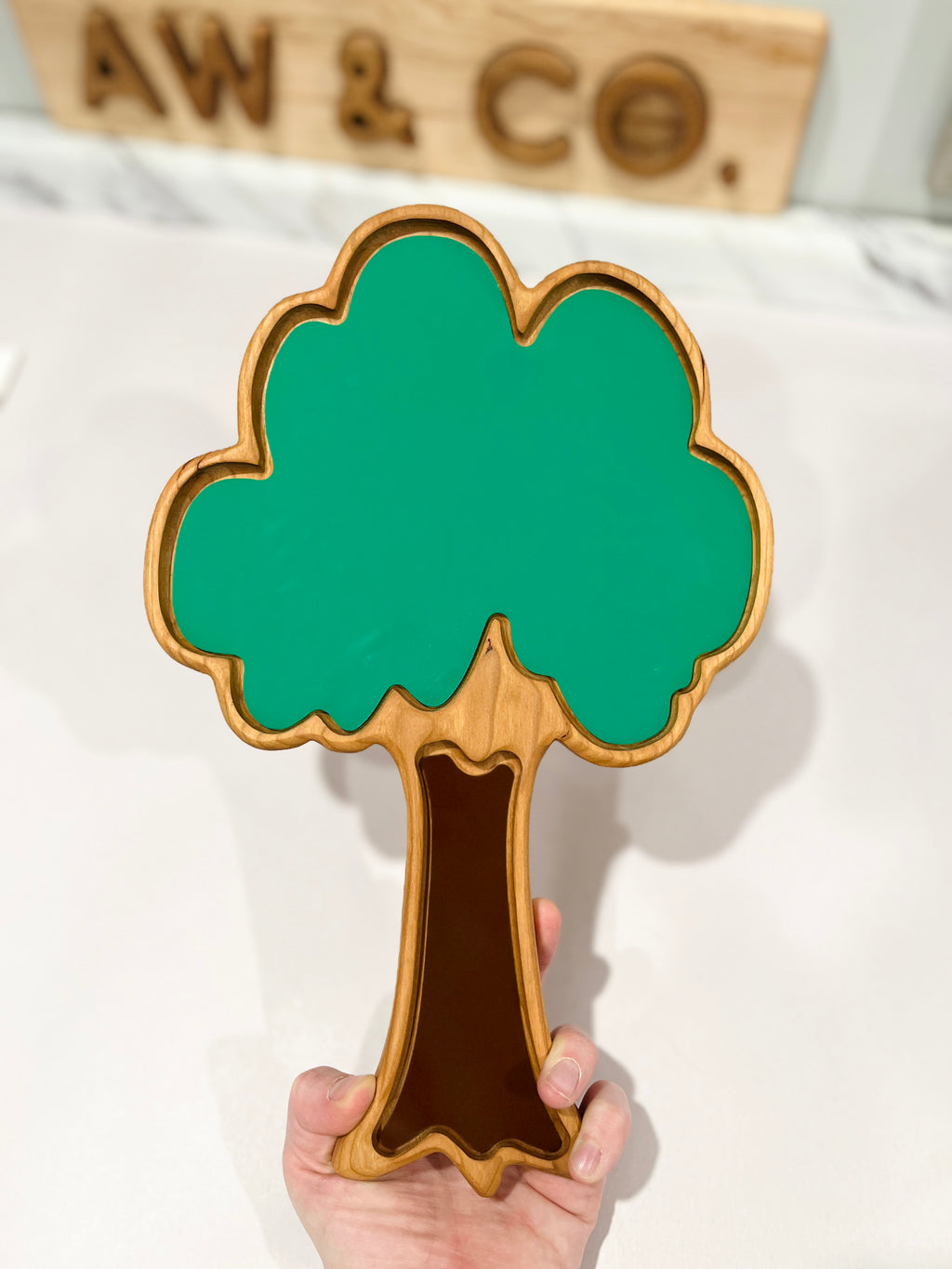 Hand holding a wooden tray shaped like a tree with green leafy top and brown trunk on a white surface