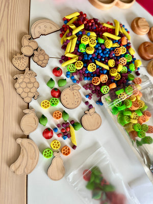 Assorted colorful plastic beads and pasta shapes mixed with wooden fruit cutouts on a white surface next to a clear plastic bag