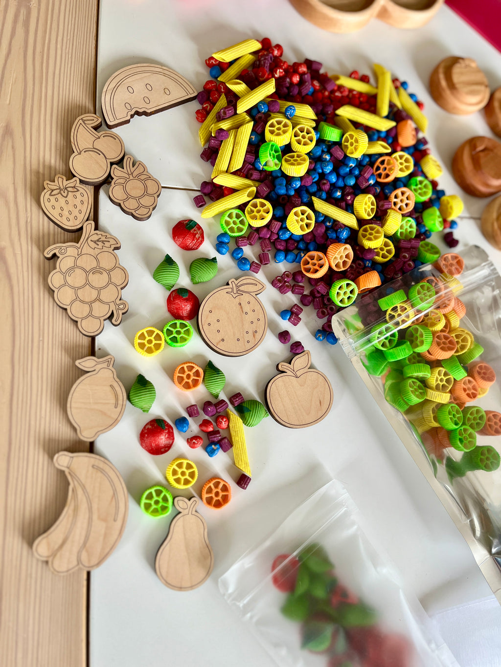 Assorted colorful plastic beads and pasta shapes mixed with wooden fruit cutouts on a white surface next to a clear plastic bag
