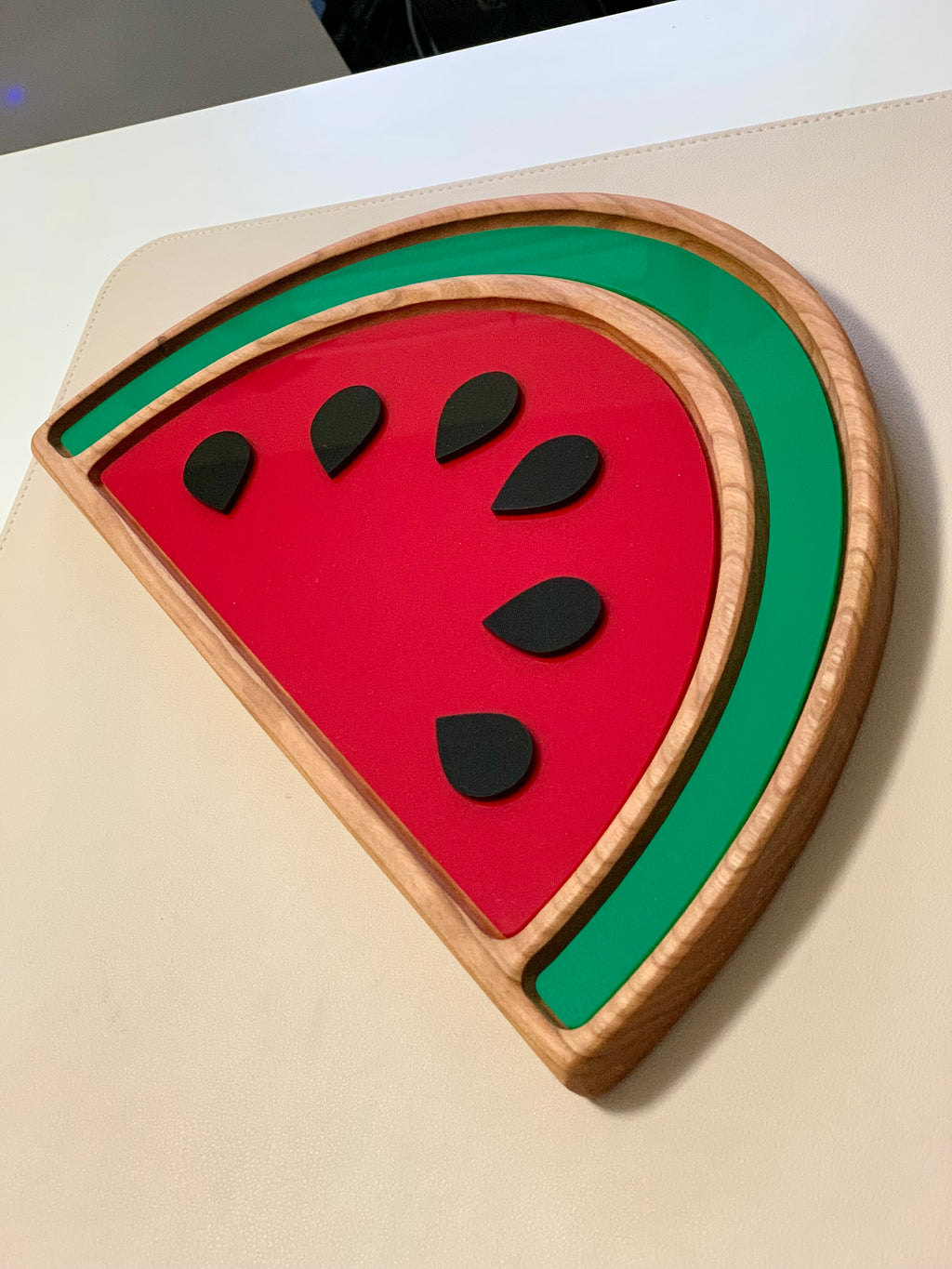 Wooden watermelon-shaped sensory tray with red and green sections and six black seed pieces on beige surface