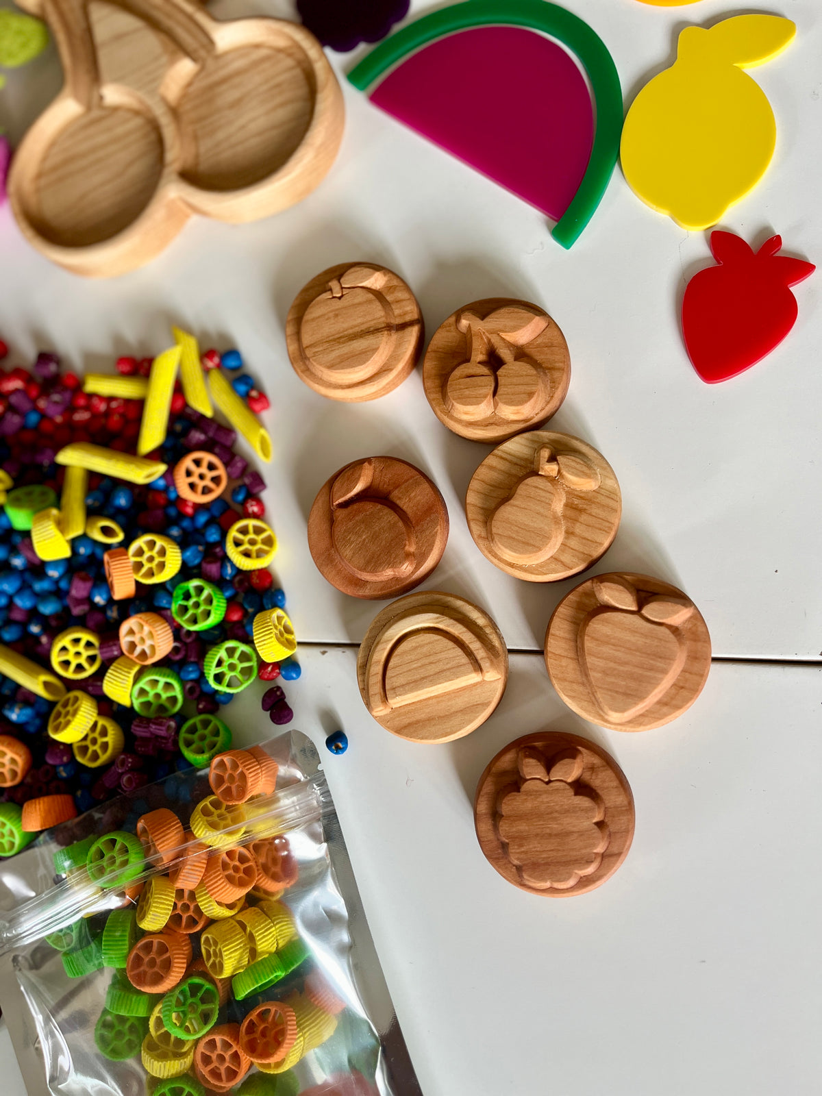 Seven round wooden fruit stamps with colorful pasta and fruit-shaped play trays on white surface