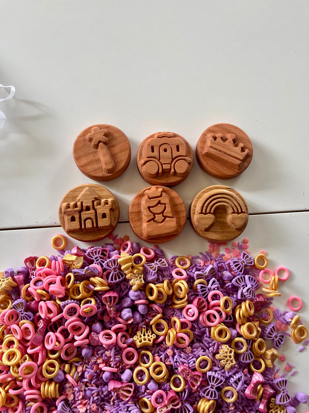 Six round wooden tokens with carved fairy tale symbols above colorful pink, purple, and yellow plastic rings and shapes on white surface