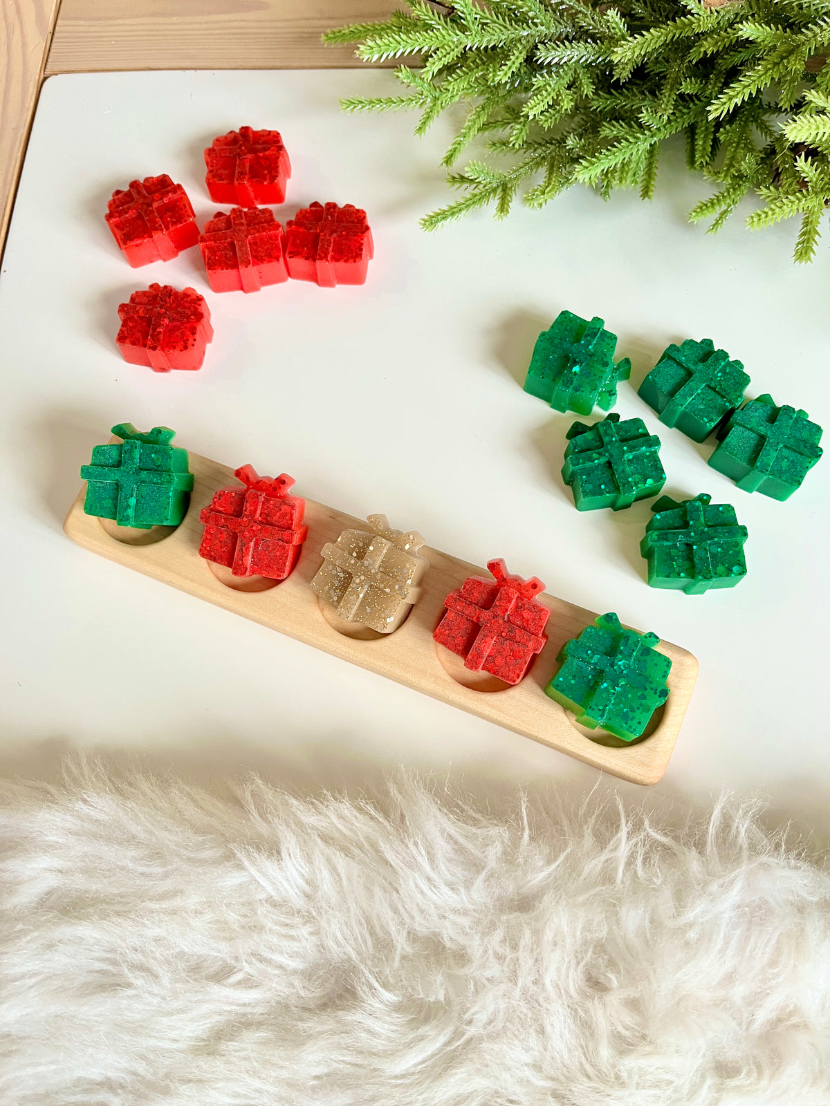 Assorted small gift-shaped wax melts in red, green, and gold resting on a wooden tray and white surface with greenery nearby