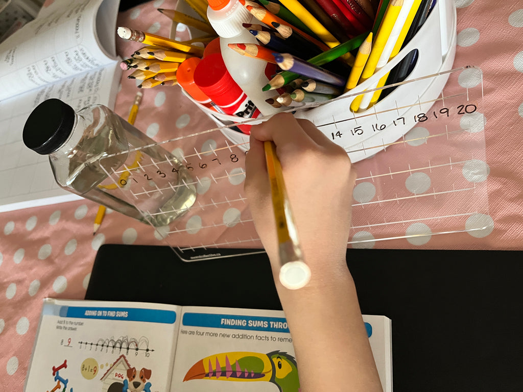 Child writing with pencil on clear acrylic number line marked 0 to 20 on pink polka dot table with colored pencils nearby