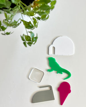 Five colorful acrylic shapes including white cave, green lizard, clear rounded square, gray rock, and pink ice cream on white surface with green plant above