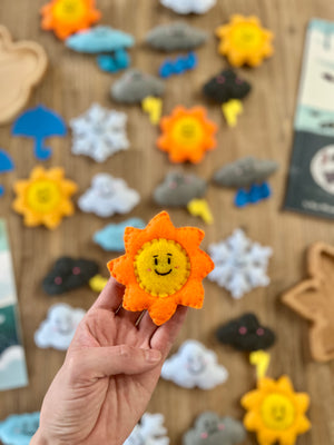 Hand holding an orange felt sun with a smiling face, surrounded by assorted colorful Felt Weather Shapes, Made in Canada, handcrafted, homeschool, education resources.