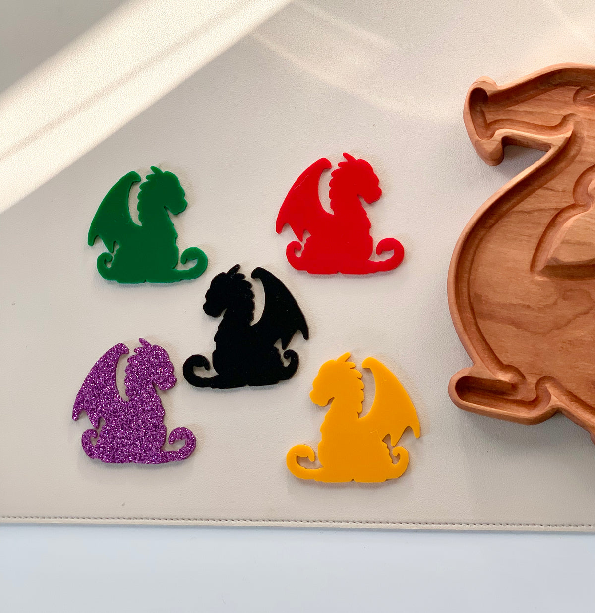 Five colorful dragon-shaped plastic pieces in green, red, black, yellow, and purple glitter next to a wooden dragon tray