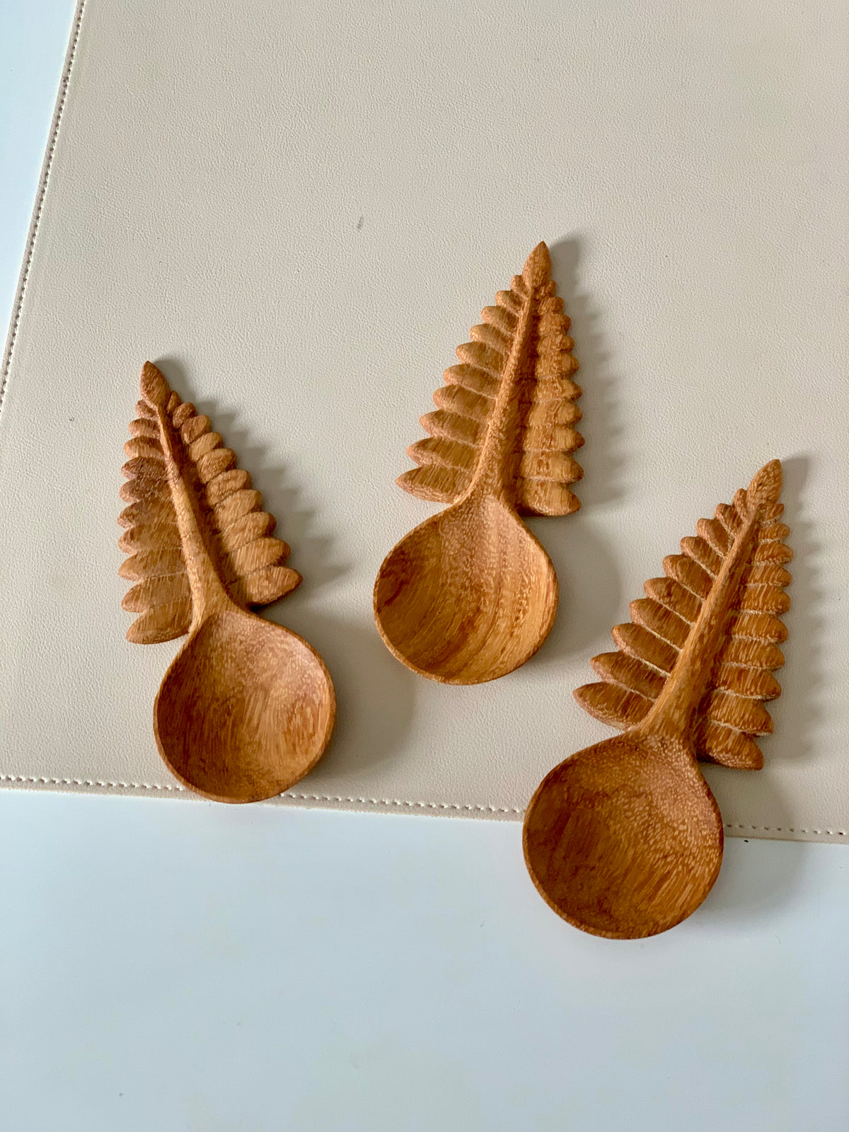 Three wooden spoons with handles carved into fern leaf shapes resting on a beige textured surface
