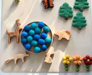 Round wooden tray filled with blue and teal felt balls surrounded by wooden animal and tree figures and felt flowers