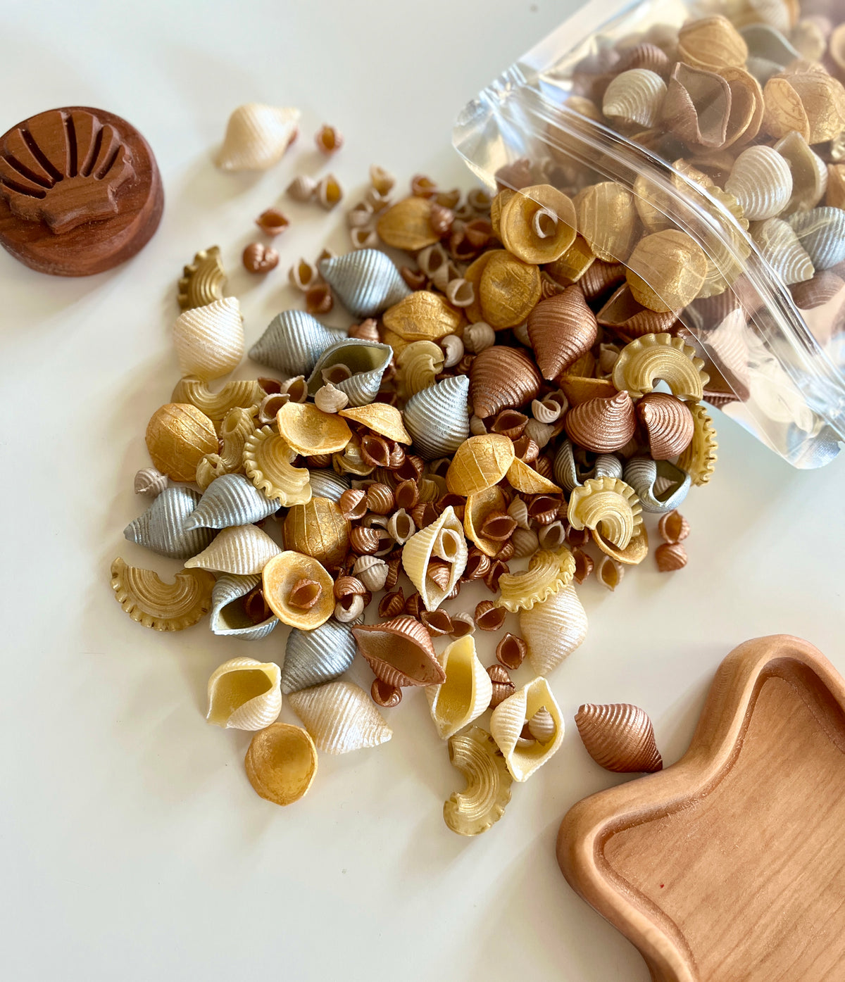 Assorted natural and metallic-colored seashells spilling from clear bag onto white surface beside wooden objects
