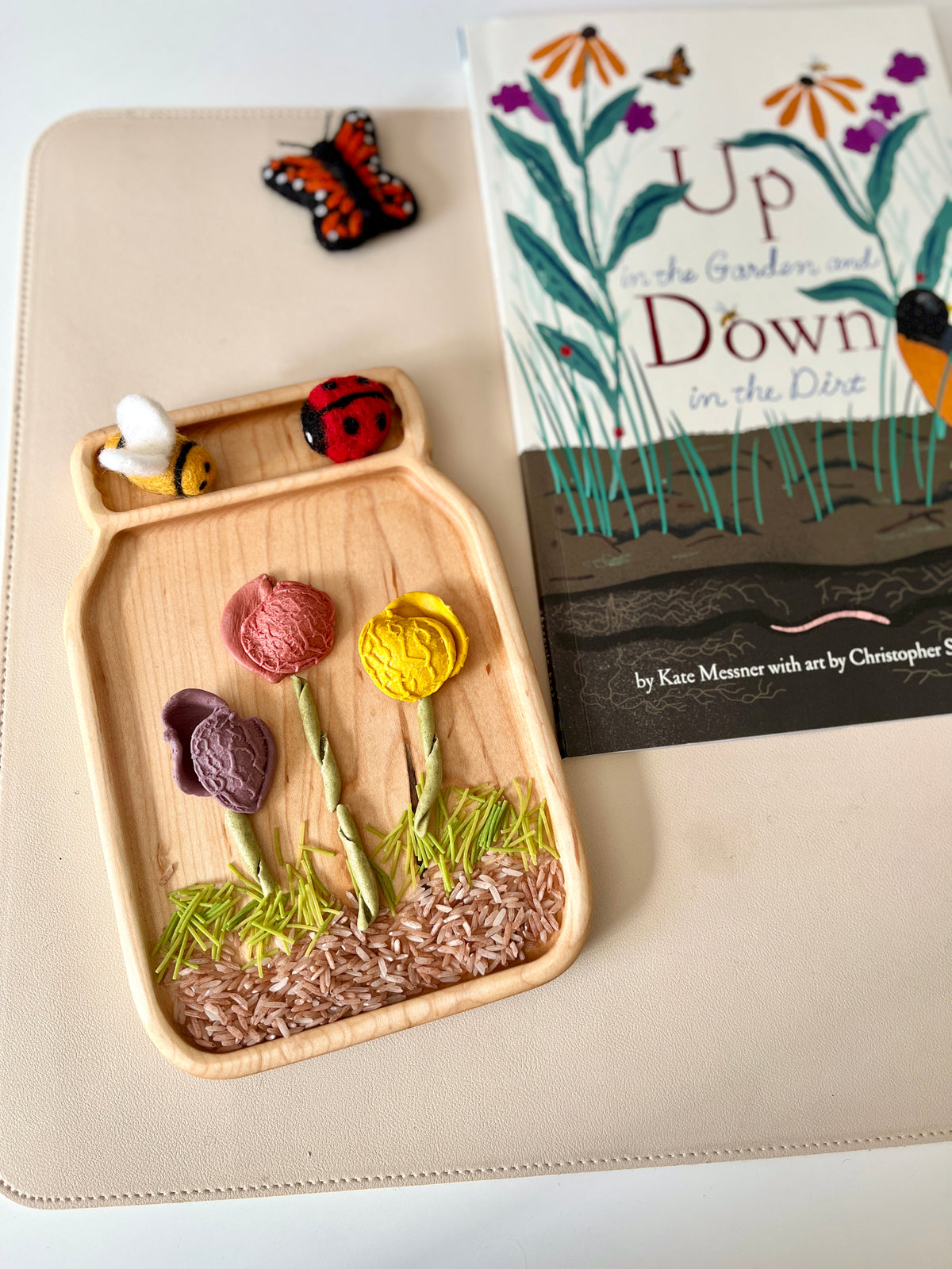 Wooden tray shaped like a jar holding fabric flowers, green grass, and felt bee and ladybug decorations nearby a book with floral cover art