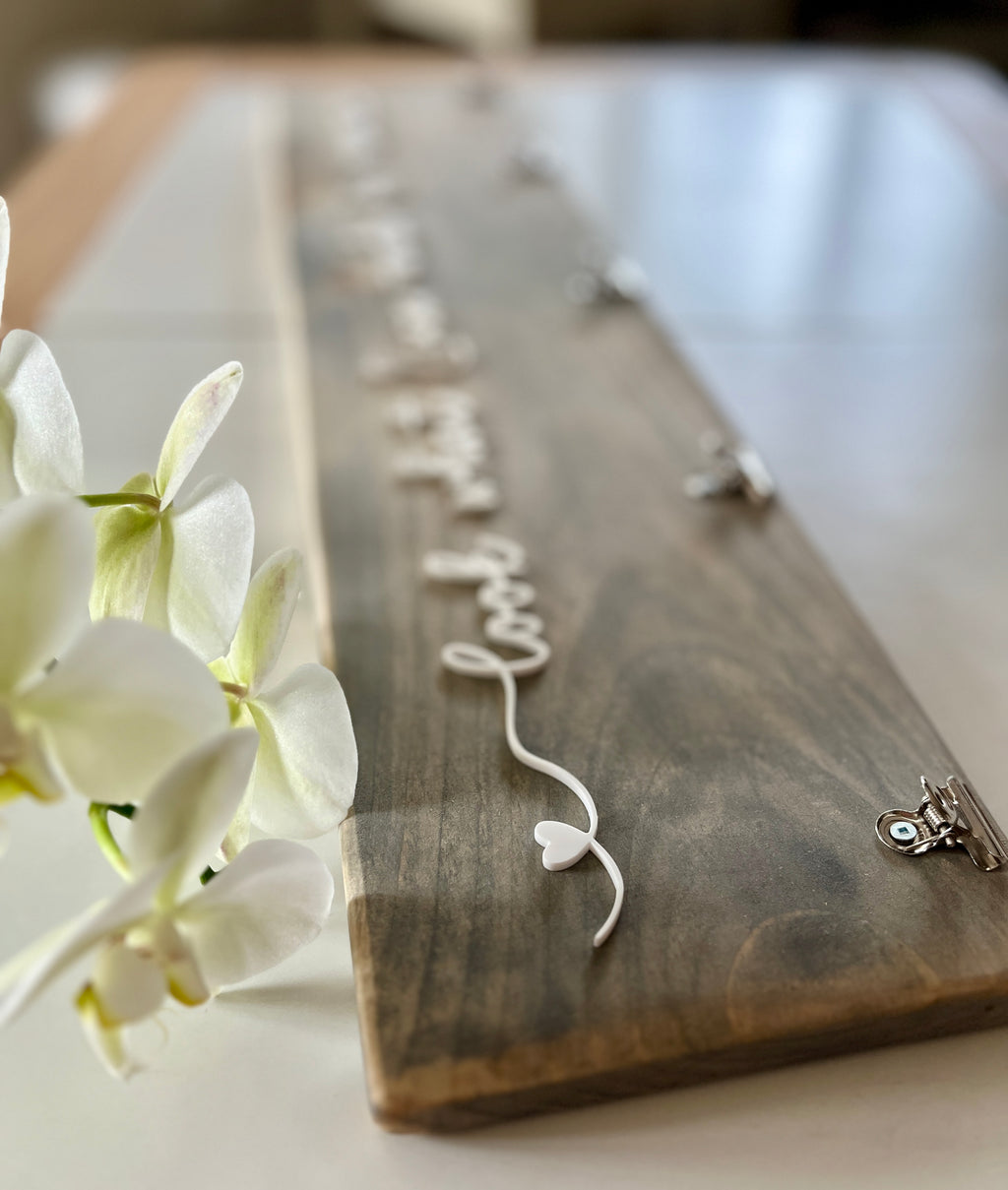 Wooden art display sign with white cursive lettering and heart detail, natural wood grain background with clip holders on white table